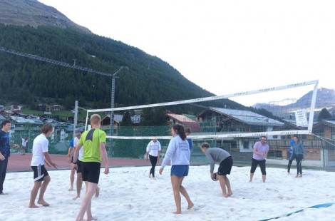 The ski team are playing volleyball at the base of the mountain. All the players are looking towards one person, who is about to serve. The mountain in the background is dark with lots of foliage.